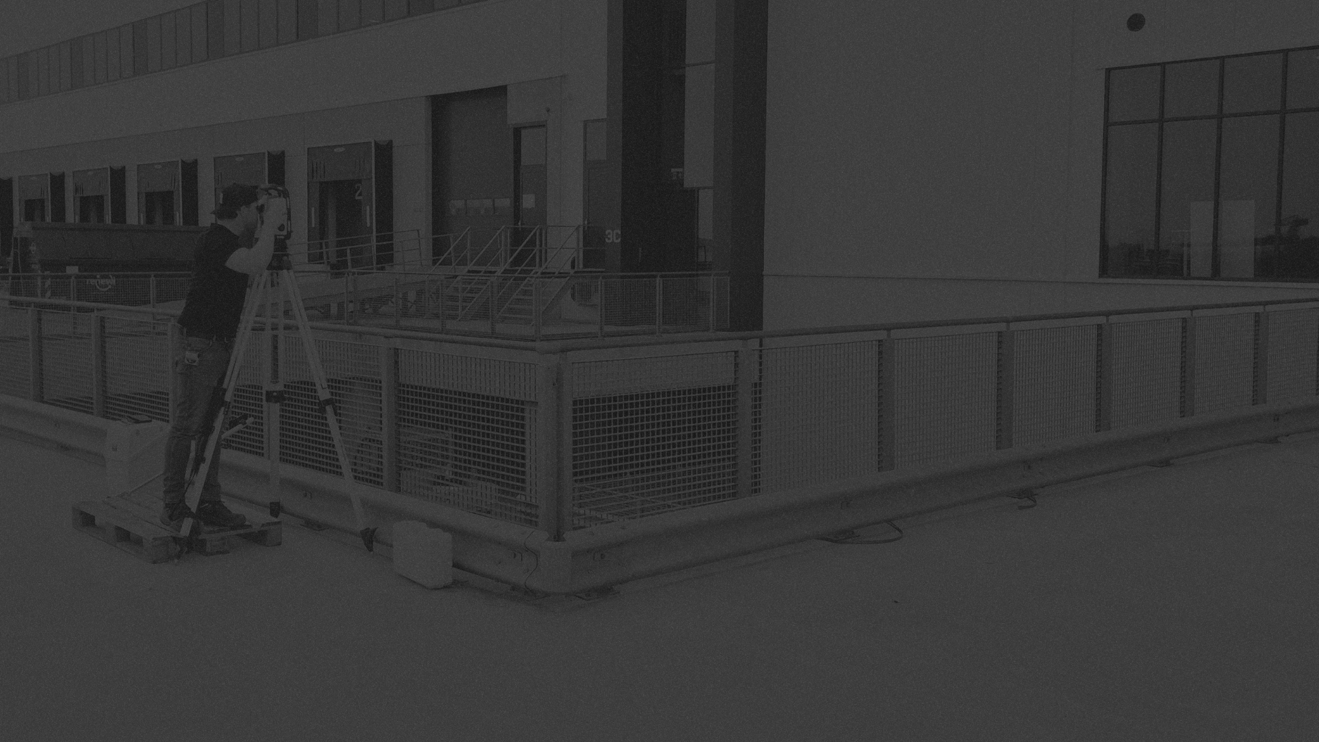 A dark, low-contrast outdoor scene showing a person standing on a ladder near a metal railing in front of a modern building.
